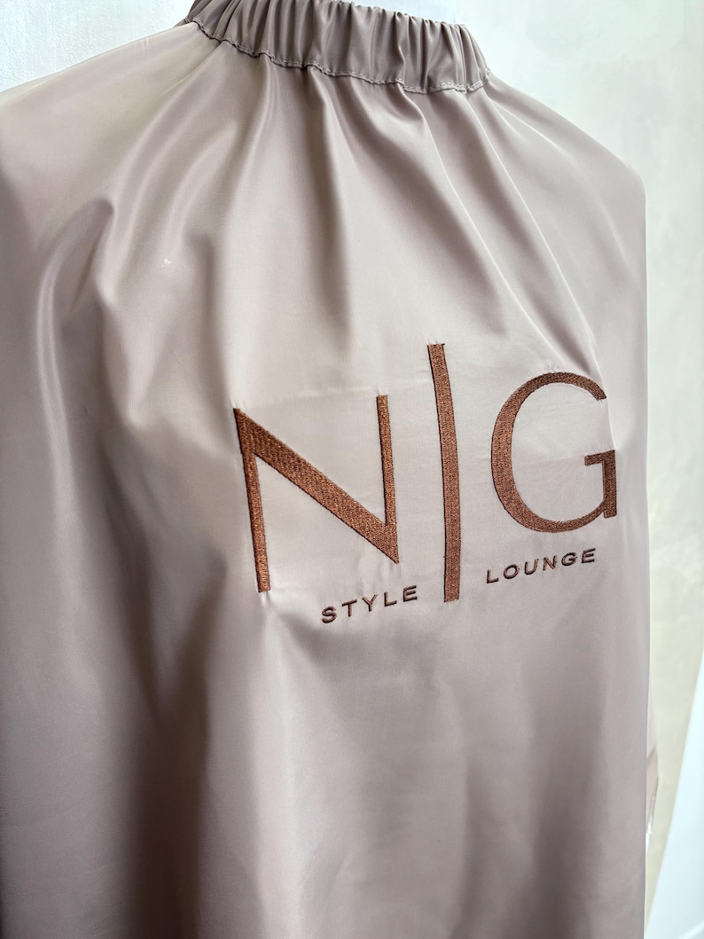 May include: A light brown hair cutting cape with the text "N|G STYLE LOUNGE" embroidered in brown on the front.