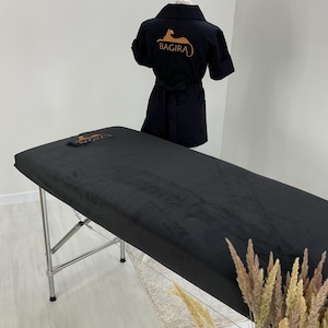 May include: A black massage table with a matching black cover and a black robe with the logo "BAGIRA" in orange. The table has a silver metal frame. The robe is displayed on a mannequin. Decorative dried plants are in the foreground.