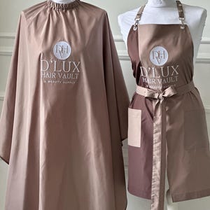 May include: A set of two brown salon capes with the logo "D'LUX HAIR VAULT & BEAUTY SUPPLY" embroidered on them. One cape is a full-length cape with a neck opening, and the other is a shorter apron-style cape with a tie.