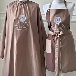 May include: A taupe hair salon cape with a gathered neck and a taupe apron featuring two front pockets and adjustable straps. Both items display a white circular logo with the text DHV D'LUX HAIR VAULT & BEAUTY SUPPLY embroidered on the front.