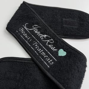 May include: Black terry cloth headband with embroidered text "Janet Rose Beauty Treatments" in white script. A small, light blue heart is next to the text. The headband has a hook and loop closure.