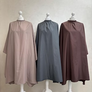 May include: Three hair cutting capes on white mannequins. The capes are made of a water-resistant material and come in beige, gray, and burgundy.