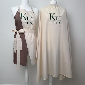 Può includere: Un grembiule marrone e crema con cintura da annodare e una mantella da parrucchiere color crema, entrambi con il logo verde scuro KK Kay's Kollection Hair Stylist stampato sul davanti. Il grembiule ha un design bicolore e la mantella un collo arricciato.