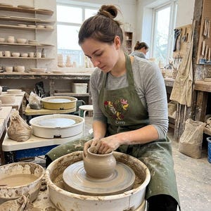 May include: An olive green apron featuring an embroidered heart-shaped floral design and the name Corey in white script. The apron is worn over a long-sleeved gray shirt while shaping a clay pot on a pottery wheel in a studio setting.