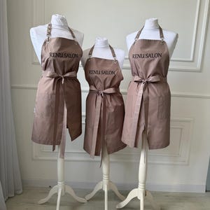 Può includere: Tre grembiuli color tortora esposti su manichini bianchi, ognuno con la scritta RENU SALON stampata in nero sul petto. I grembiuli presentano lacci regolabili al collo con fibbie in metallo, un laccio in vita e due tasche frontali inferiori.