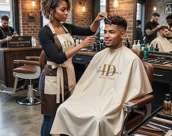 May include: A barber giving a haircut to a customer in a salon. The barber is wearing a brown and beige apron with the logo "ID's Joanna Danai's BEAUTY BAR". The customer is wearing a beige cape with the same logo. Other barbers are working in the background.