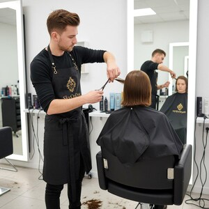 Può includere: Un parrucchiere con un grembiule nero con la scritta "Sky Or Dive" taglia i capelli di un cliente con le forbici in un salone. Il cliente è seduto su una sedia nera, con un mantello nero. Uno specchio riflette un altro parrucchiere e cliente. Il salone ha un'illuminazione brillante.