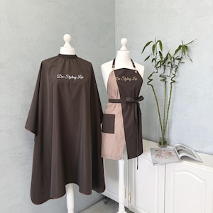 May include: A dark brown salon cape with an elastic neck and the white embroidered text Da Styling Lab, displayed next to a two-tone dark brown and beige apron with a waist tie, a front pocket, and the white embroidered text Da Styling Lab.