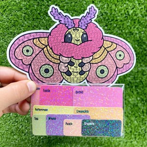Op de afbeelding: Een kleurrijke sticker van een mot met een roze, gele en groene mozaïekpatroon. De sticker is op een witte achtergrond en wordt geleverd met een vel gekleurde papierstalen met namen zoals "Orchid", "Buttercream", "Thee" en "Grafiet".
