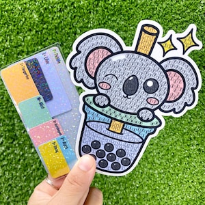 Puede incluir: Un koala gris sosteniendo una taza de té de boba con una pajita. El koala está hecho de pequeñas cuentas de colores. El té de boba está hecho de pequeñas cuentas negras. El fondo es verde.