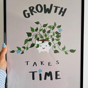 May include: A pink framed print with a white pot with a smiling face and a green plant growing out of it. The text "GROWTH TAKES TIME" is written in black letters.