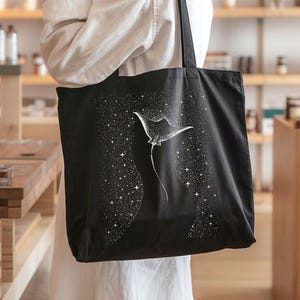 May include: Black tote bag featuring a white illustration of a manta ray against a starry background. The bag has long handles and is made of a sturdy fabric. The design is simple and elegant, suitable for everyday use.