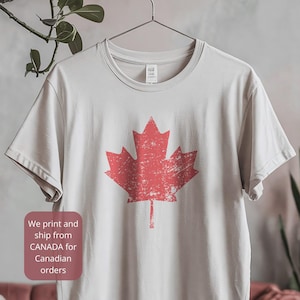 May include: A light beige short-sleeved t-shirt featuring a large, distressed red maple leaf graphic centered on the chest. The garment is displayed on a hanger, showcasing a crew neckline and a standard fit with a clean, minimalist aesthetic.