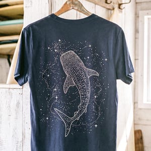 Puede incluir: Una camiseta de manga corta en color azul marino oscuro con un gráfico de líneas blancas en la espalda. El diseño muestra un tiburón ballena nadando entre un campo de estrellas dispersas y constelaciones celestiales, con un estilo limpio.