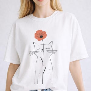 May include: White t-shirt featuring a minimalist cat illustration with a red flower. The cat is drawn with simple black lines, and the flower has a dark center. The shirt is a casual, oversized fit.
