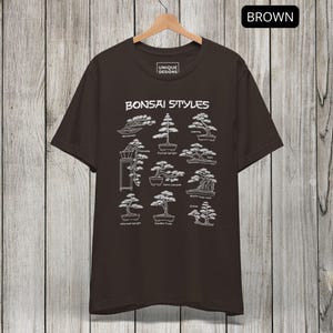 May include: A dark brown short-sleeve t-shirt featuring a white graphic print titled Bonsai Styles. The design displays nine distinct bonsai tree illustrations with labels including Windswept, Cascade, Formal Upright, Leaning, Raft, Semi-cascade, Root-over-rock, Informal Upright, Double Trunk, and Group.