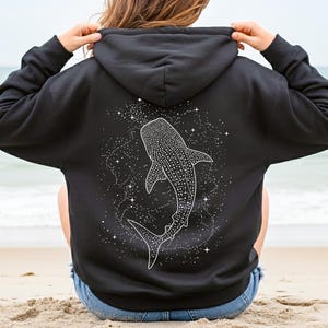 May include: Black hoodie featuring a white illustration of a whale shark surrounded by stars. The design is on the back of the hoodie. The person is sitting on a sandy beach with the ocean in the background.