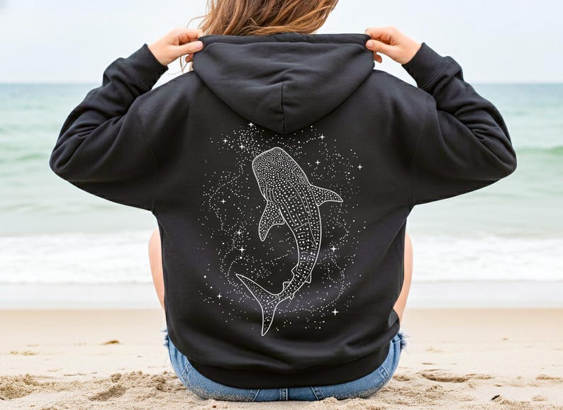 May include: Black hoodie featuring a white illustration of a whale shark surrounded by stars. The design is on the back of the hoodie. The person is sitting on a sandy beach with the ocean in the background.