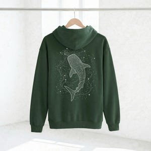 May include: A dark green hoodie with a white whale shark design on the back. The whale shark is surrounded by a constellation of white stars. The hoodie has a hood and long sleeves and is hanging on a wooden hanger.