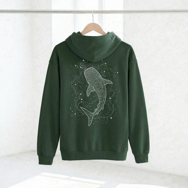 May include: A dark green hoodie with a white whale shark design on the back. The whale shark is surrounded by a constellation of white stars. The hoodie has a hood and long sleeves and is hanging on a wooden hanger.
