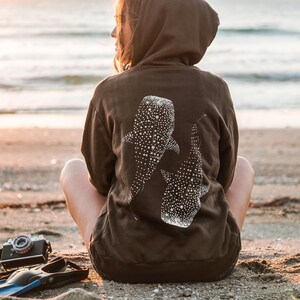 May include: A dark brown hooded sweatshirt featuring a white graphic print of two whale sharks on the back. The design displays the distinct spotted pattern of the whale sharks, with the hoodie worn by an individual facing away from the camera.
