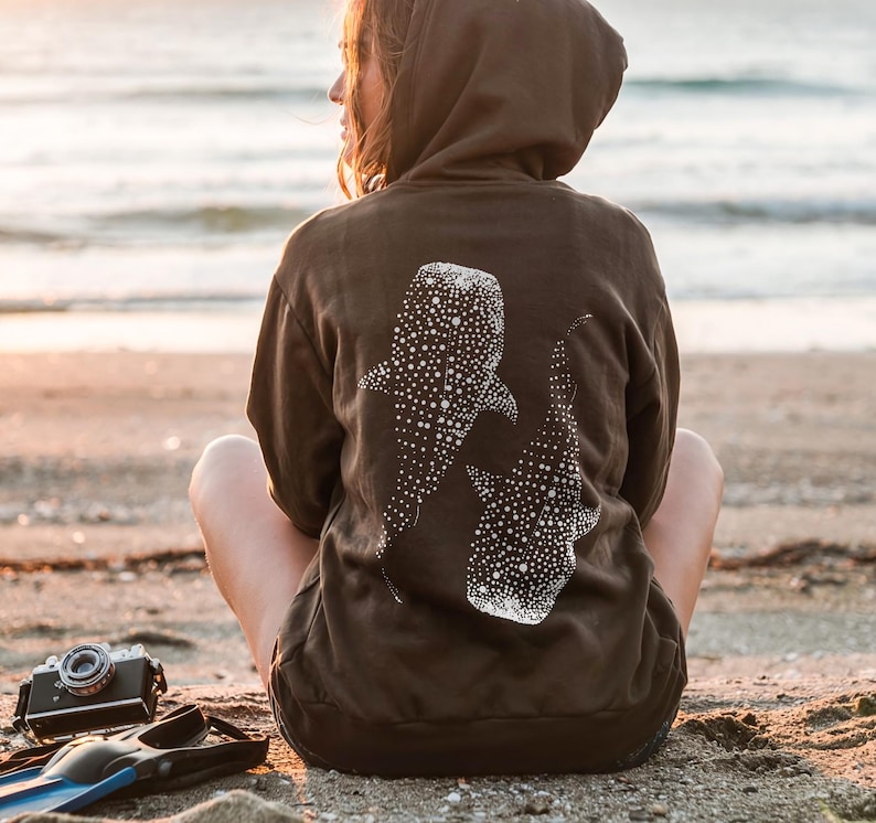 May include: A dark brown hooded sweatshirt featuring a white graphic print of two whale sharks on the back. The design displays the distinct spotted pattern of the whale sharks, with the hoodie worn by an individual facing away from the camera.