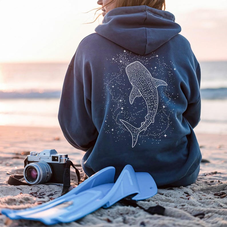 May include: A blue hoodie featuring a white whale shark design on the back, surrounded by stars. The person is sitting on a sandy beach with a vintage camera and blue flippers nearby. The hoodie is a muted blue color.
