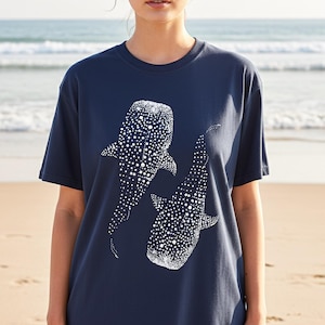 May include: Navy blue t-shirt featuring a white graphic of two whale sharks. The design is detailed with white dots, creating a textured appearance. The shirt is a casual, short-sleeved style, suitable for everyday wear.