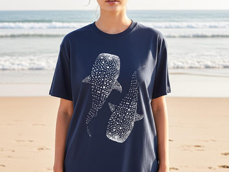May include: Navy blue t-shirt featuring a white graphic of two whale sharks. The design is detailed with white dots, creating a textured appearance. The shirt is a casual, short-sleeved style, suitable for everyday wear.