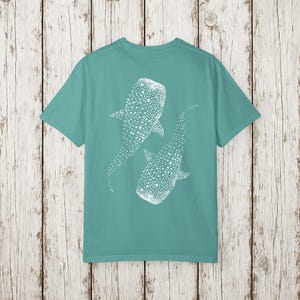 May include: Teal t-shirt featuring a white graphic of two whale sharks. The whale sharks are depicted with dotted patterns. The shirt has a crew neck and short sleeves. The background is a light wood pattern.