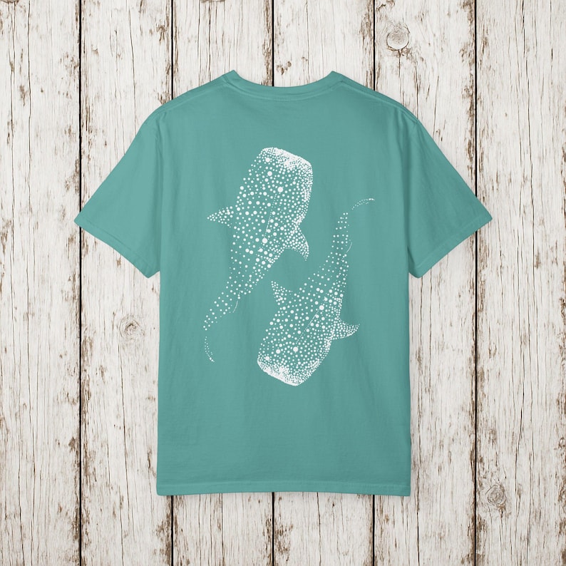 May include: Teal t-shirt featuring a white graphic of two whale sharks. The whale sharks are depicted with dotted patterns. The shirt has a crew neck and short sleeves. The background is a light wood pattern.