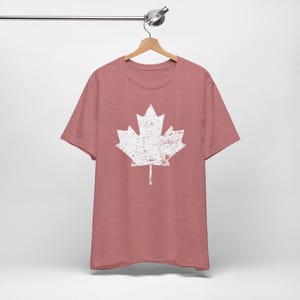Pode incluir: Uma t-shirt de manga curta em malva mesclado com um gr&aacute;fico grande e centralizado de uma folha de &aacute;cer branca. O design da folha de &aacute;cer tem uma textura desgastada e envelhecida. A t-shirt est&aacute; pendurada num cabide de madeira.