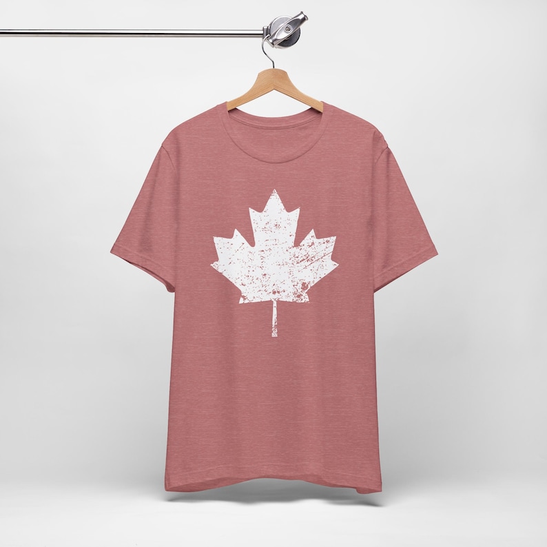 Pode incluir: Uma t-shirt de manga curta em malva mesclado com um gr&aacute;fico grande e centralizado de uma folha de &aacute;cer branca. O design da folha de &aacute;cer tem uma textura desgastada e envelhecida. A t-shirt est&aacute; pendurada num cabide de madeira.