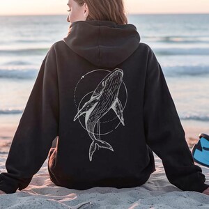 K&ouml;nnte beinhalten: Schwarzer Kapuzenpullover mit einer wei&szlig;en Wal-Illustration in einem kreisf&ouml;rmigen Design auf dem R&uuml;cken. Der Hoodie hat eine Kapuze mit Kordelzug und lange &Auml;rmel. Das Bild ist an einem Sandstrand mit dem Meer im Hintergrund aufgenommen.