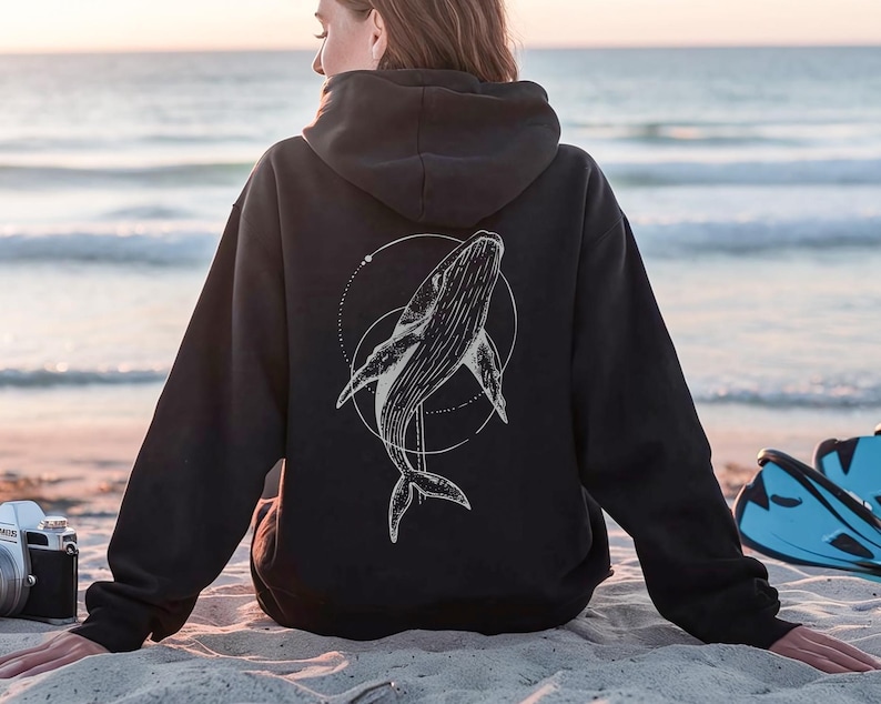 K&ouml;nnte beinhalten: Schwarzer Kapuzenpullover mit einer wei&szlig;en Wal-Illustration in einem kreisf&ouml;rmigen Design auf dem R&uuml;cken. Der Hoodie hat eine Kapuze mit Kordelzug und lange &Auml;rmel. Das Bild ist an einem Sandstrand mit dem Meer im Hintergrund aufgenommen.