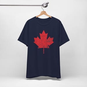 Pode incluir: Uma t-shirt azul-marinho de manga curta com um grande gr&aacute;fico central de uma folha de &aacute;cer vermelha. O design da folha de &aacute;cer tem uma textura desgastada e envelhecida. A t-shirt &eacute; apresentada num cabide de madeira sobre um fundo liso.