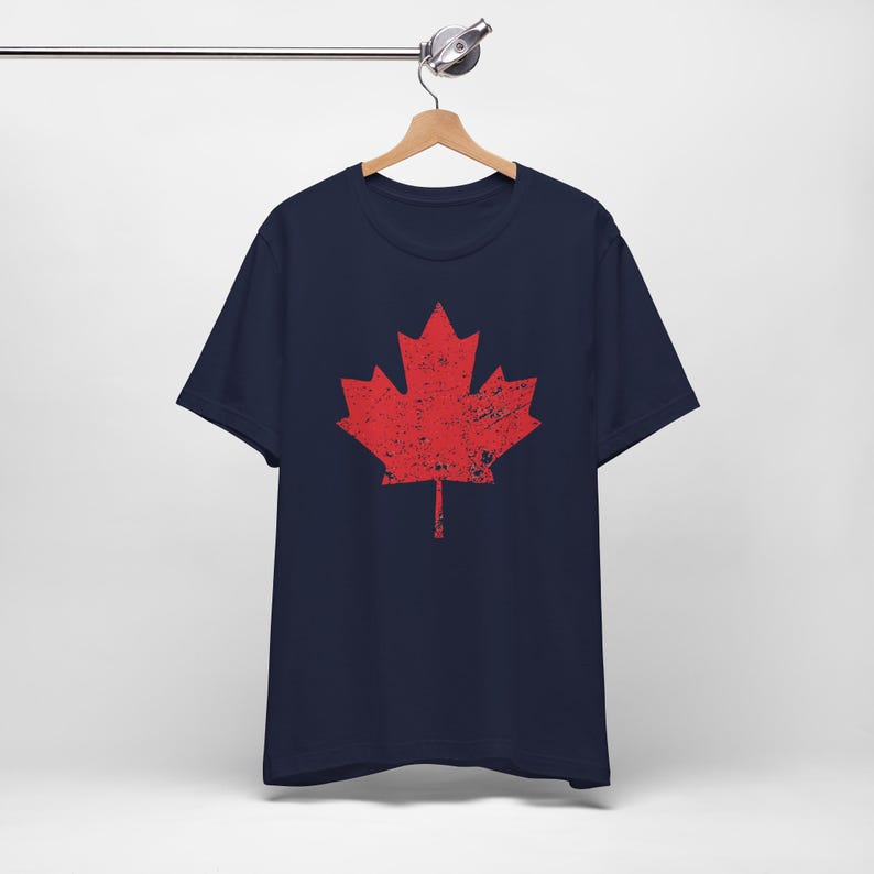 Pode incluir: Uma t-shirt azul-marinho de manga curta com um grande gr&aacute;fico central de uma folha de &aacute;cer vermelha. O design da folha de &aacute;cer tem uma textura desgastada e envelhecida. A t-shirt &eacute; apresentada num cabide de madeira sobre um fundo liso.