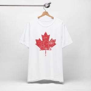 Pode incluir: Uma t-shirt branca de manga curta com um gr&aacute;fico grande e centralizado de uma folha de &aacute;cer vermelha. O design da folha de &aacute;cer apresenta uma textura desgastada e envelhecida em toda a superf&iacute;cie. A t-shirt est&aacute; num cabide de madeira.
