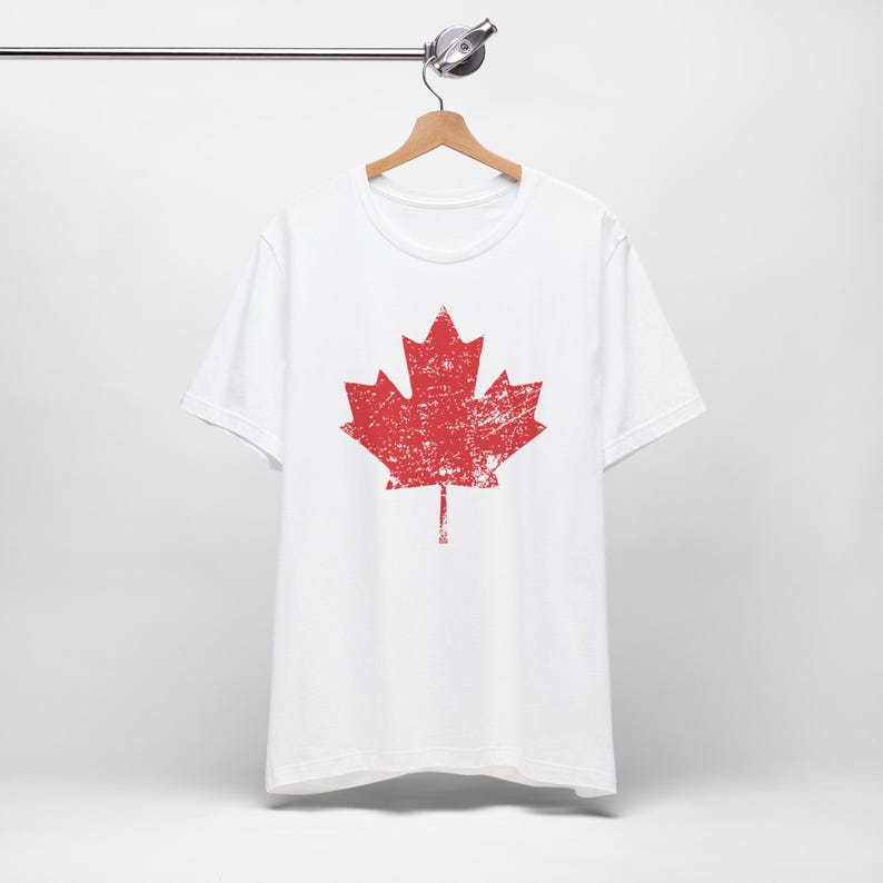 Pode incluir: Uma t-shirt branca de manga curta com um gr&aacute;fico grande e centralizado de uma folha de &aacute;cer vermelha. O design da folha de &aacute;cer apresenta uma textura desgastada e envelhecida em toda a superf&iacute;cie. A t-shirt est&aacute; num cabide de madeira.