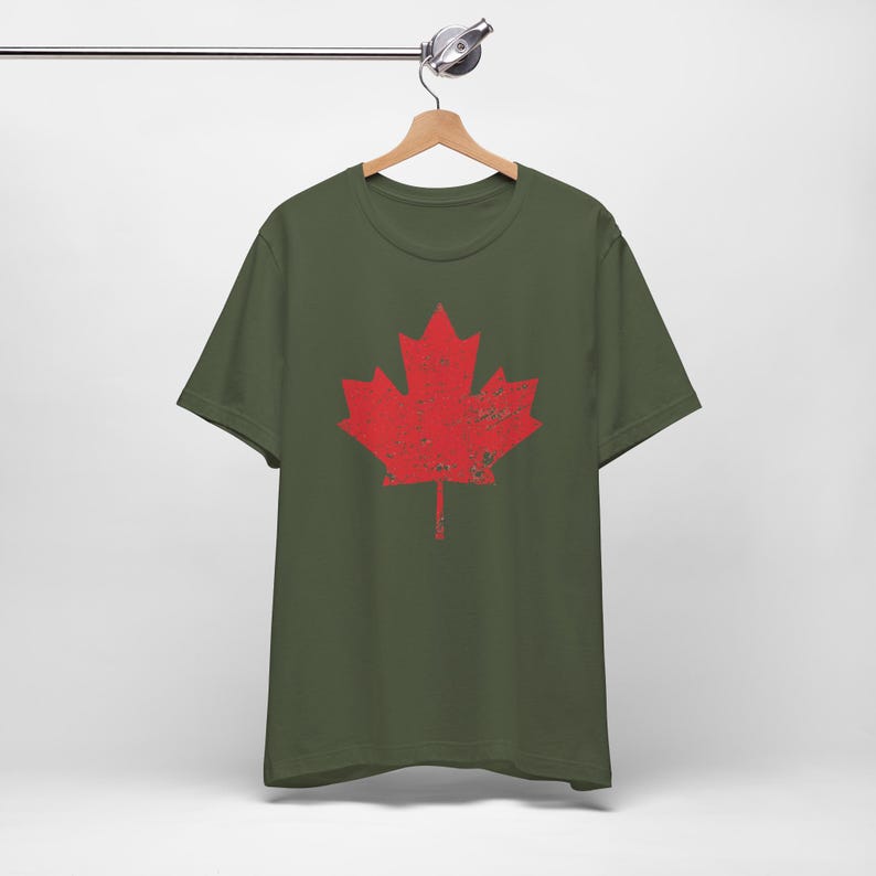 Pode incluir: Uma t-shirt de manga curta verde-azeitona com um gr&aacute;fico grande de uma folha de &aacute;cer vermelha ao centro. O design da folha de &aacute;cer tem uma textura desgastada e envelhecida em toda a superf&iacute;cie. A t-shirt tem gola redonda e est&aacute; num cabide.