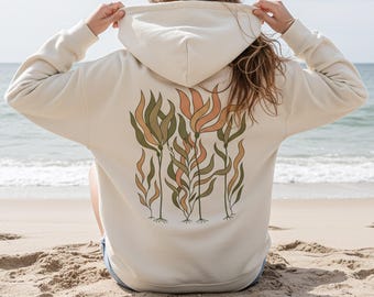 May include: Cream-colored hoodie with a floral design on the back. The design features stylized plants in shades of green, orange, and brown. The hoodie is worn on a sandy beach with the ocean in the background.