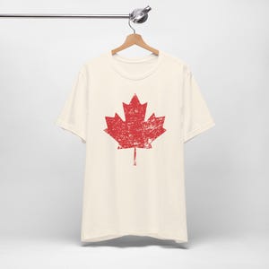 Pode incluir: Uma t-shirt de manga curta na cor creme com um gr&aacute;fico grande de uma folha de &aacute;cer vermelha ao centro. O design da folha de &aacute;cer tem uma textura desgastada e envelhecida, conferindo-lhe um aspeto usado. A t-shirt est&aacute; num cabide de madeira.