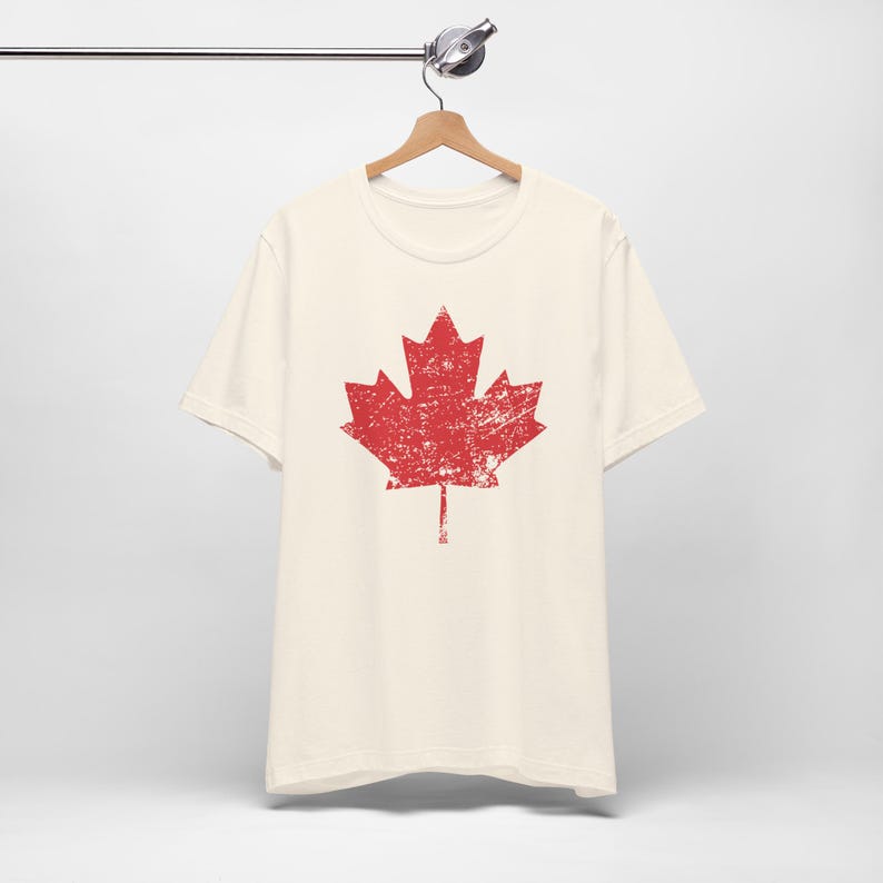 Pode incluir: Uma t-shirt de manga curta na cor creme com um gr&aacute;fico grande de uma folha de &aacute;cer vermelha ao centro. O design da folha de &aacute;cer tem uma textura desgastada e envelhecida, conferindo-lhe um aspeto usado. A t-shirt est&aacute; num cabide de madeira.
