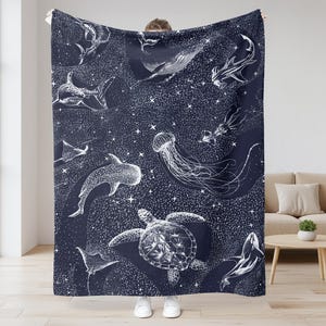 May include: A navy blue blanket featuring a white illustration of various sea creatures, including whales, sharks, and jellyfish. The design includes a starry background, creating a marine theme. The blanket is held up by a person.