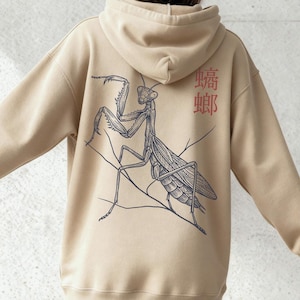 Pode incluir: Moletom bege com uma ilustração detalhada em preto e cinza de um louva-a-deus nas costas. O moletom também apresenta caracteres japoneses vermelhos. Uma peça de roupa confortável e casual.