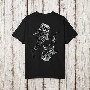 May include: Black t-shirt featuring a white graphic of two whale sharks. The whale sharks are depicted with a dotted pattern, creating a textured effect. The design is centered on the back of the shirt.