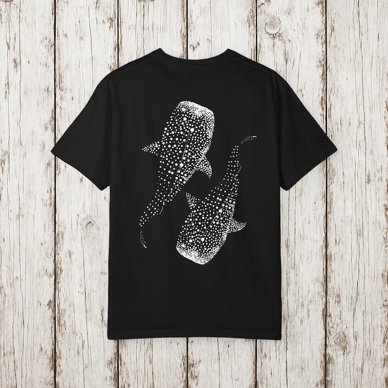 May include: Black t-shirt featuring a white graphic of two whale sharks. The whale sharks are depicted with a dotted pattern, creating a textured effect. The design is centered on the back of the shirt.