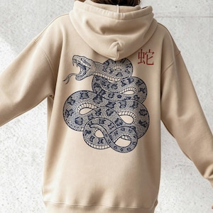 Könnte beinhalten: Beigefarbener Kapuzenpullover mit einer detaillierten Illustration einer zusammengerollten Schlange in Grau- und Schwarztönen. Das japanische Schriftzeichen für Schlange ist rot aufgedruckt. Der Hoodie hat eine Kapuze und lange Ärmel.