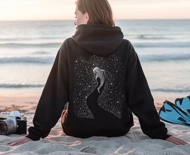 Peut inclure: Un sweat &agrave; capuche noir avec une illustration de baleine sur le dos, sur fond de nuit &eacute;toil&eacute;e. Le sweat &agrave; capuche est port&eacute; sur une plage avec un appareil photo et des palmes &agrave; proximit&eacute;. La baleine est blanche et les &eacute;toiles sont blanches.