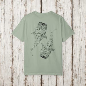 May include: Sage green t-shirt featuring a black illustration of two whale sharks. The whale sharks are depicted with dotted patterns. The t-shirt has short sleeves and a crew neck.